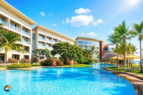 Resort-style hotel with a large blue swimming pool, palm trees, and sun loungers under yellow umbrellas on a sunny day.