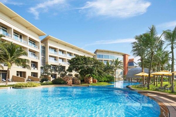 Resort-style hotel with a large blue swimming pool, palm trees, and sun loungers under yellow umbrellas on a sunny day.